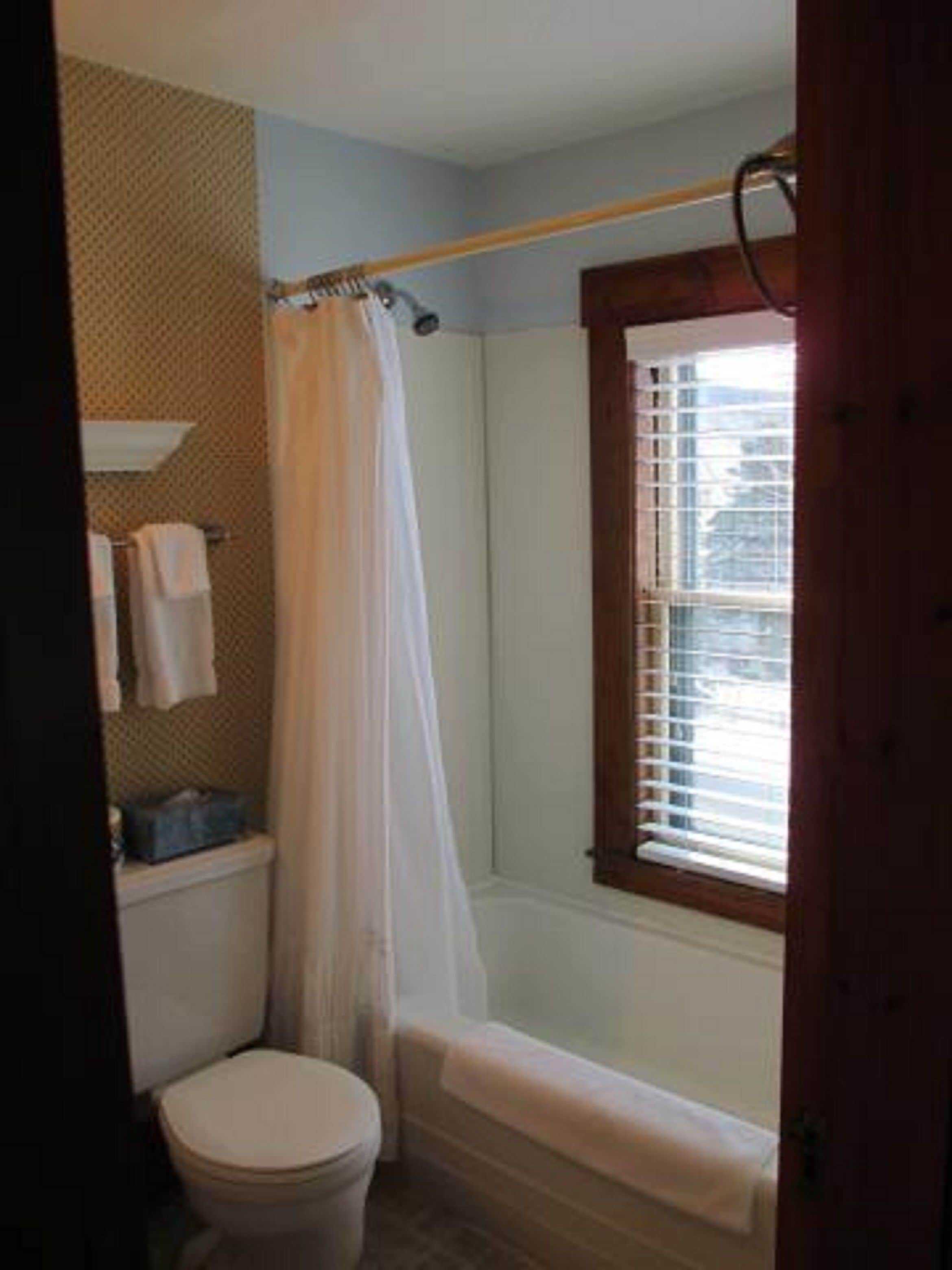 A bathroom with a toilet, a shower with a curtain, a window with blinds, and a towel rack with towels.