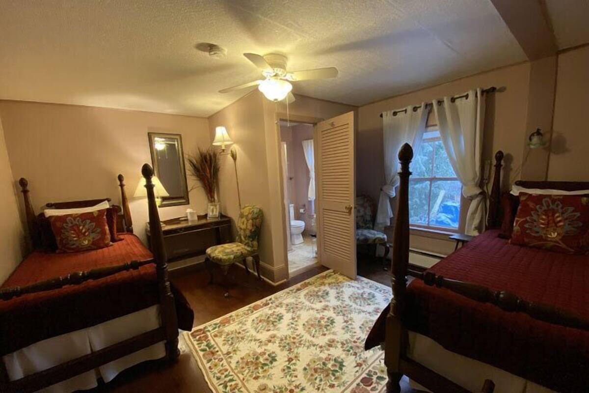 A bedroom with two beds, a ceiling fan, a large window with curtains, a bedside table, and a chair.