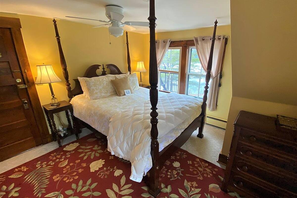 A four-poster bed with a canopy, a wooden floor, a red floral carpet, a wooden dresser, and a window with curtains.