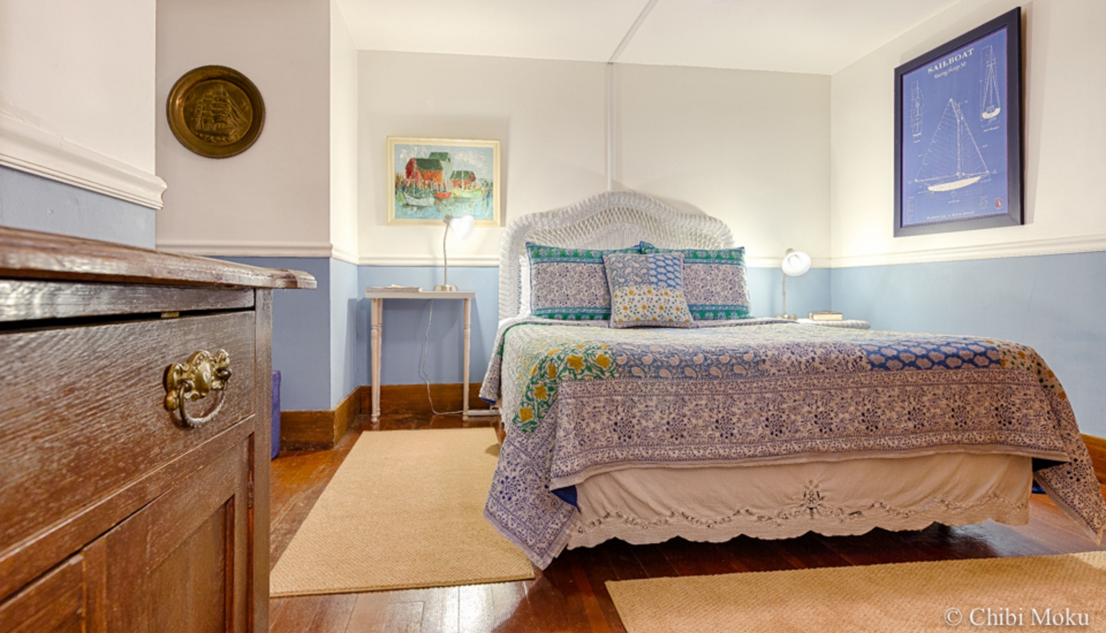 A bedroom with a wooden dresser, a bed with patterned bedding, a framed picture on the wall, and a nautical-themed poster.