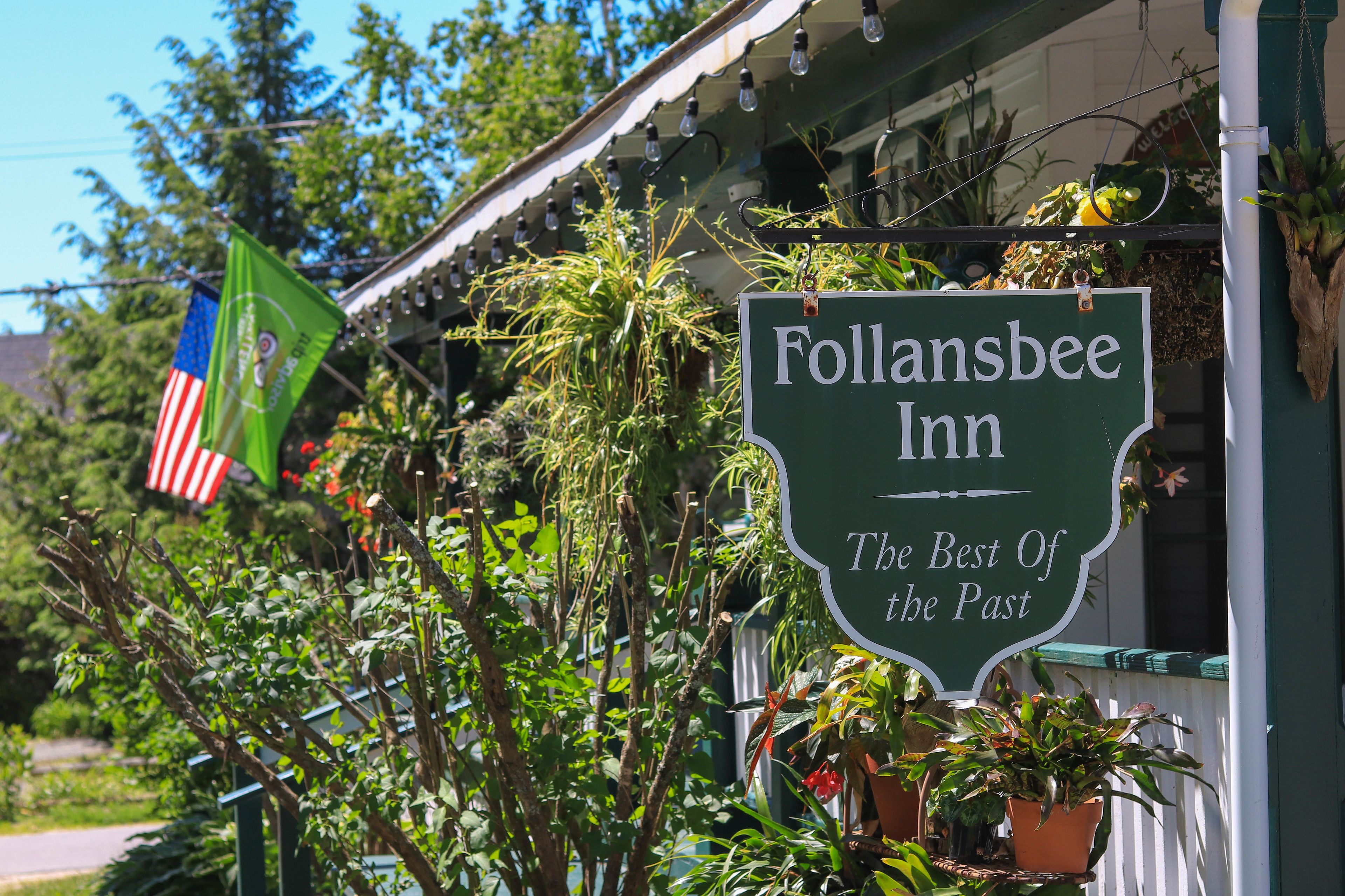 A sign for Follansbee Inn with a green background and white lettering, surrounded by greenery and hanging plants.
