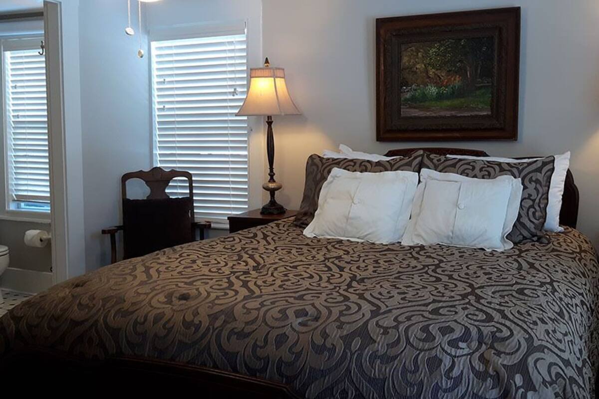 A bedroom with a patterned bedspread, two bedside lamps, a painting on the wall, and a window with blinds.