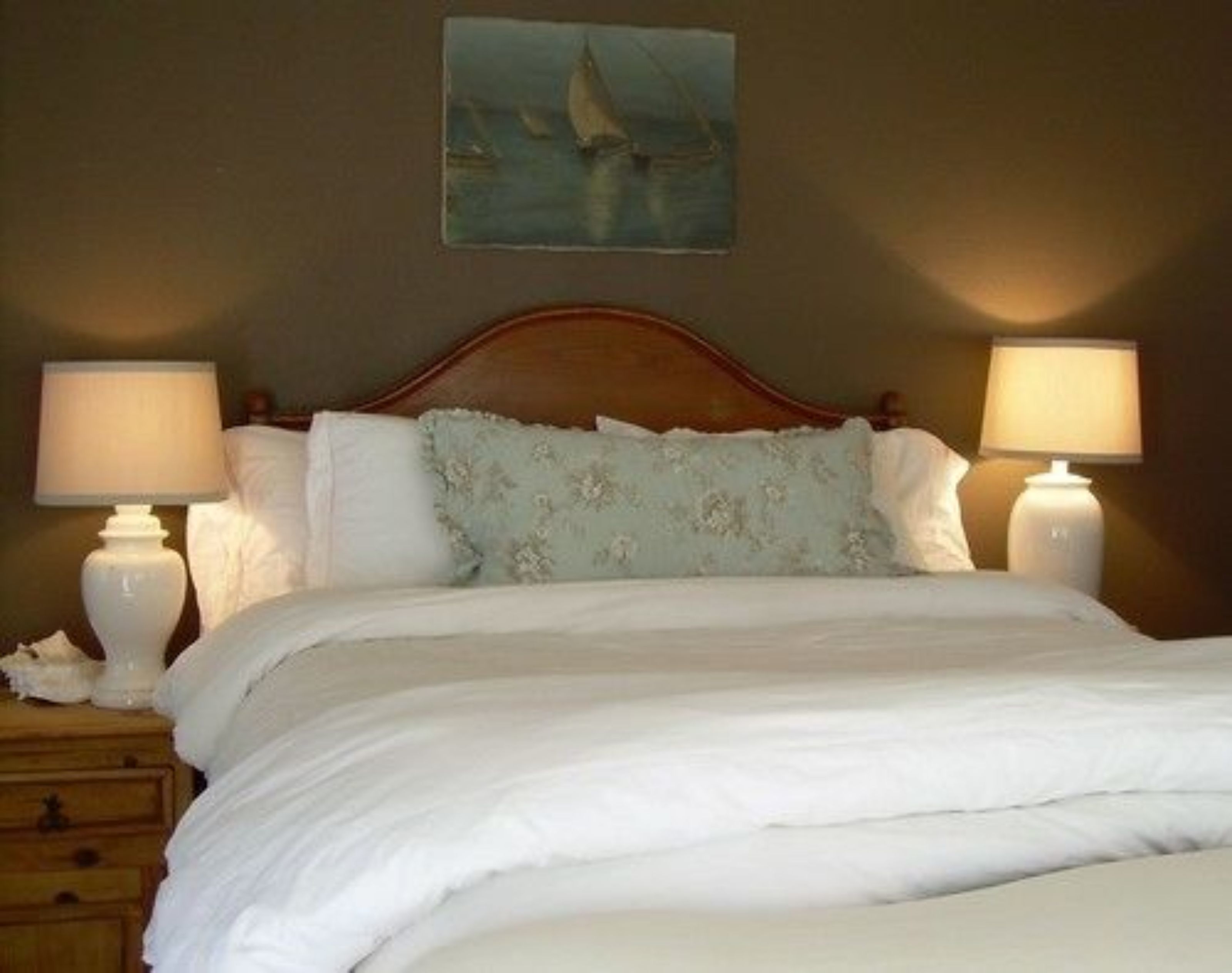 Uma cama bem arrumada com roupa de cama branca, duas luminárias de cabeceira e uma pintura de um veleiro na parede.