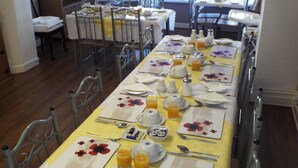 Uma área de refeições com mesas preparadas para o pequeno-almoço, com toalhas de mesa florais e sumo de laranja.