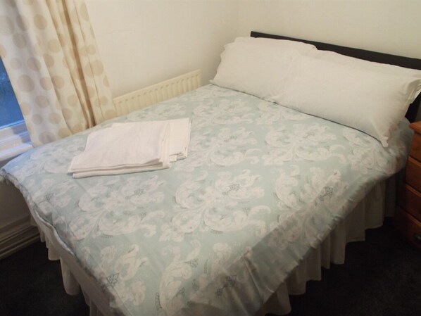 A neatly made bed with a floral bedspread and white pillows, a wooden nightstand, and a window with curtains.