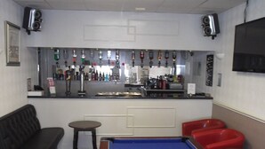 Uma área de bar com mesa de bilhar, cadeiras vermelhas, sofá preto e balcão com diversos garrafas e tela de televisão.