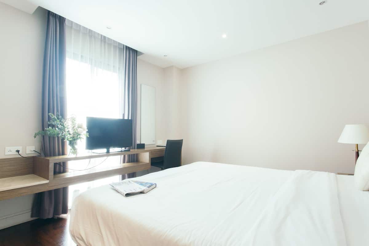 Ein Hotelzimmer mit einem großen Bett, einem Schreibtisch, einem Sessel, einem Fernseher, einer Lampe und einem Fenster mit Vorhängen.