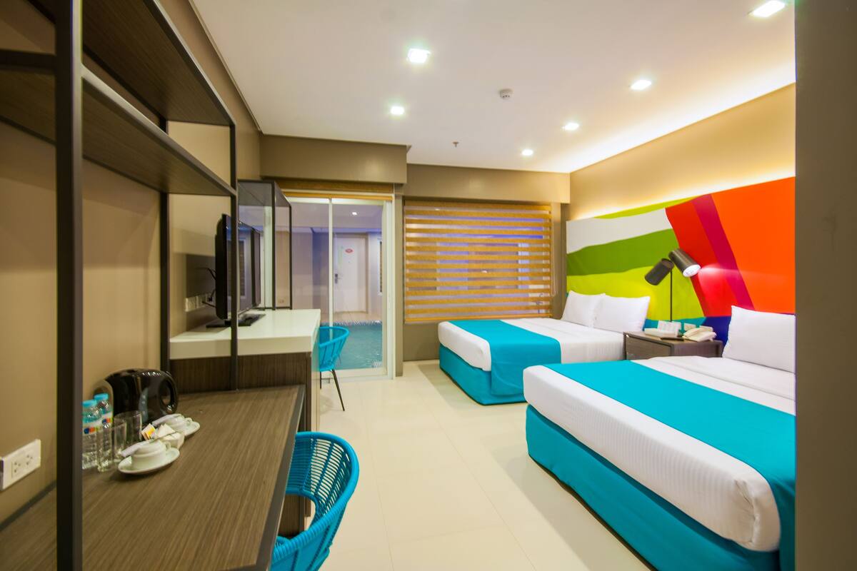 Habitación de hotel con dos camas, un escritorio con una silla, una mesita con una tetera y una ventana con persianas.
