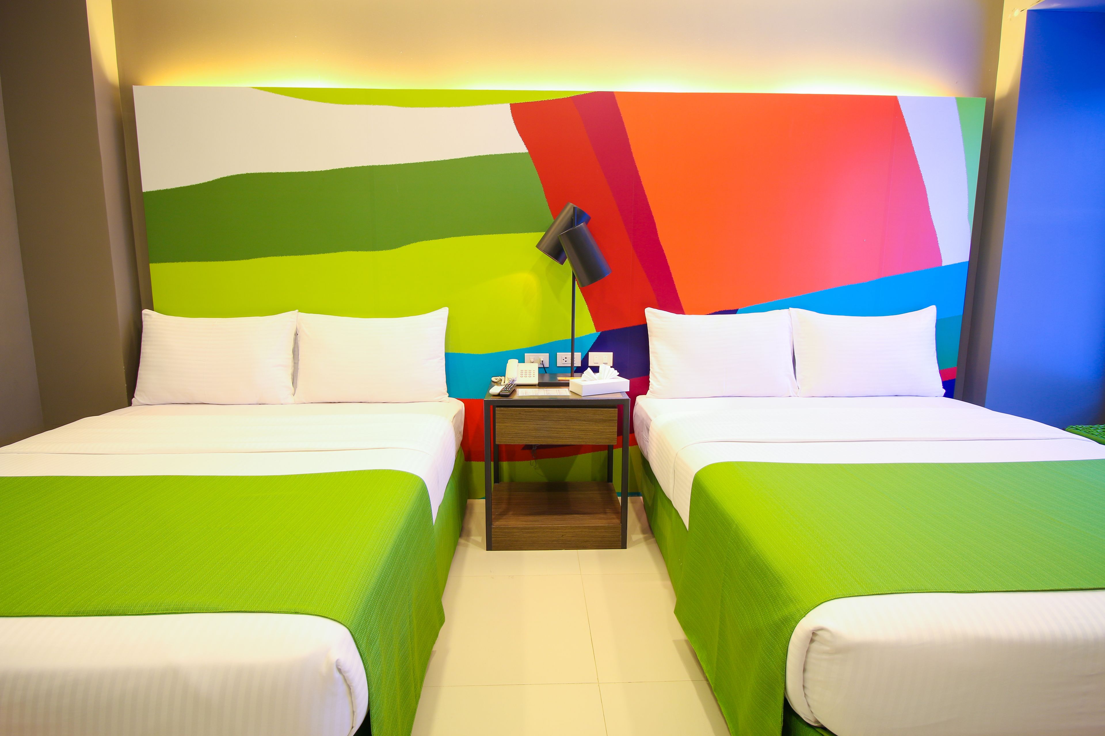 Habitación de hotel con dos camas, cada una con ropa de cama verde, una mesita de noche con lámpara y un diseño abstracto y colorido en la pared.