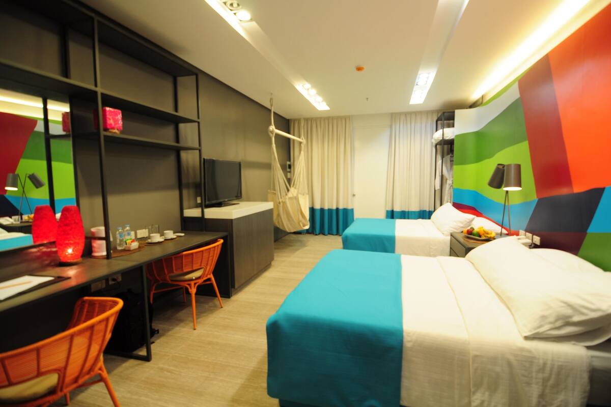 Una habitación de hotel con dos camas, un escritorio con silla, un televisor y una pared colorida.