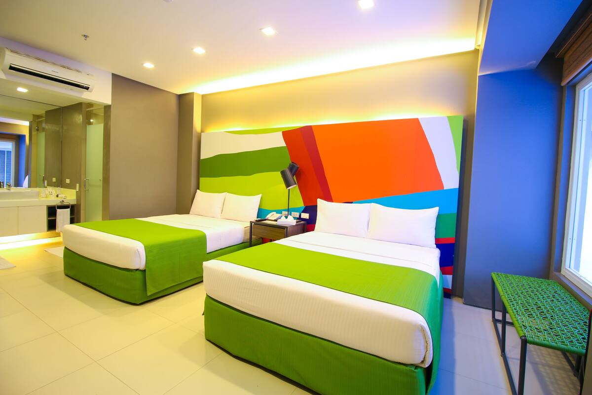 Una habitación de hotel con dos camas, una mesita, una mesita de noche y un baño con espejo y lavamanos.