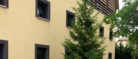 A building with a beige facade, multiple windows, and a well-maintained garden with a large coniferous tree and a flower bed.