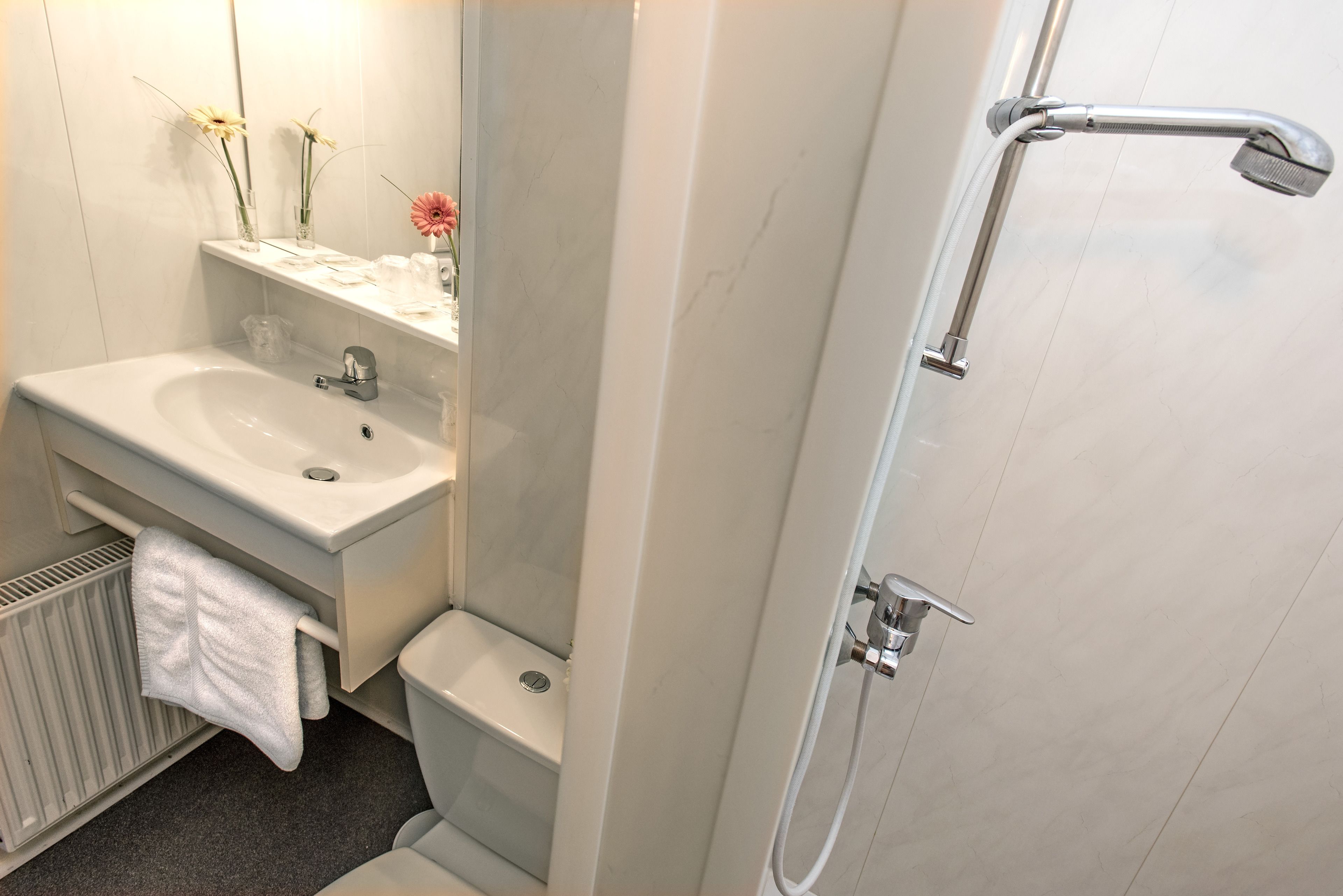 Un bany amb un lavabo, vàter i dutxa de color blanc. Hi ha una tovallola penjada al radiador, i un mirall sobre el lavabo.