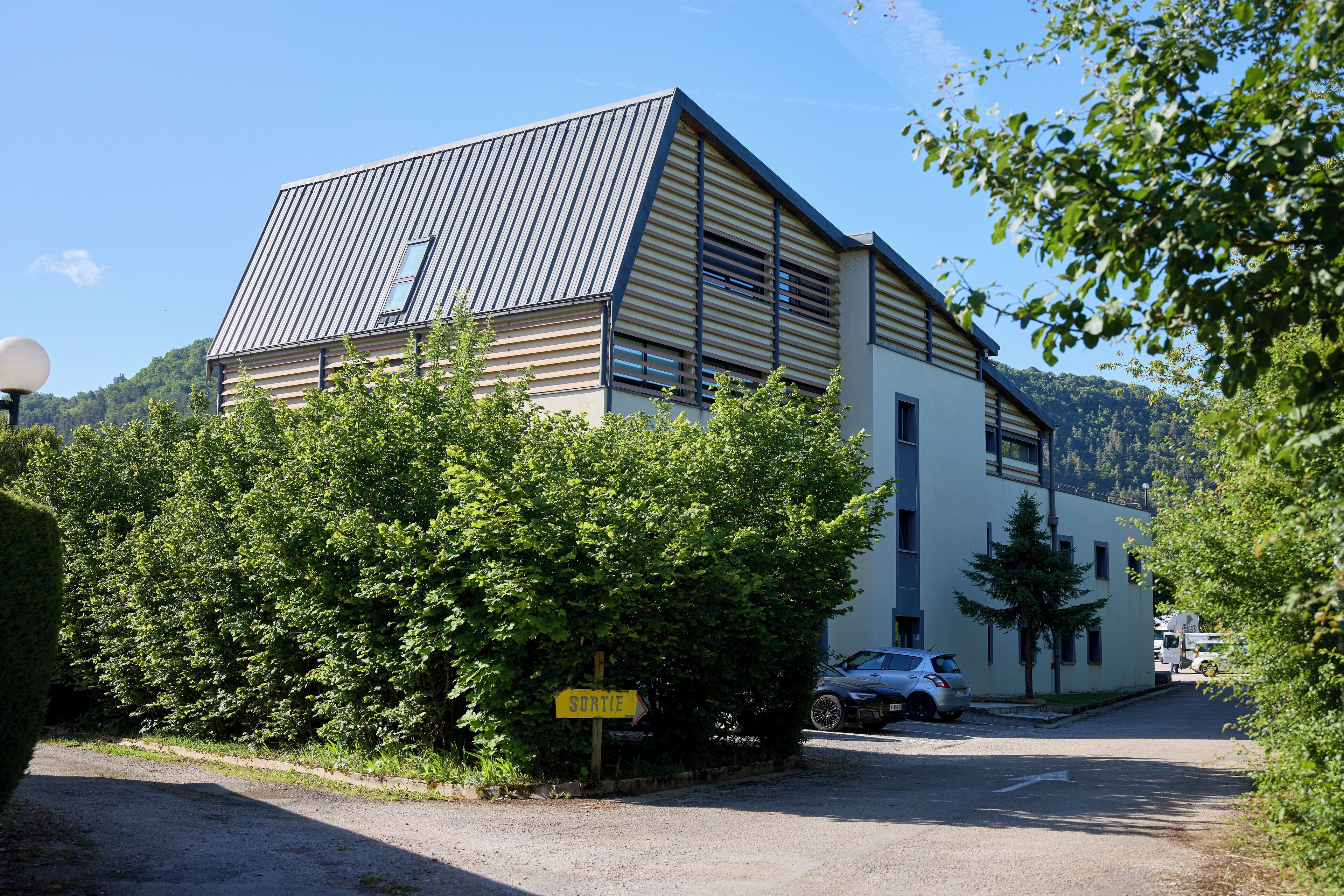 Een modern gebouw met een schuin dak, omgeven door groen en een heldere lucht.