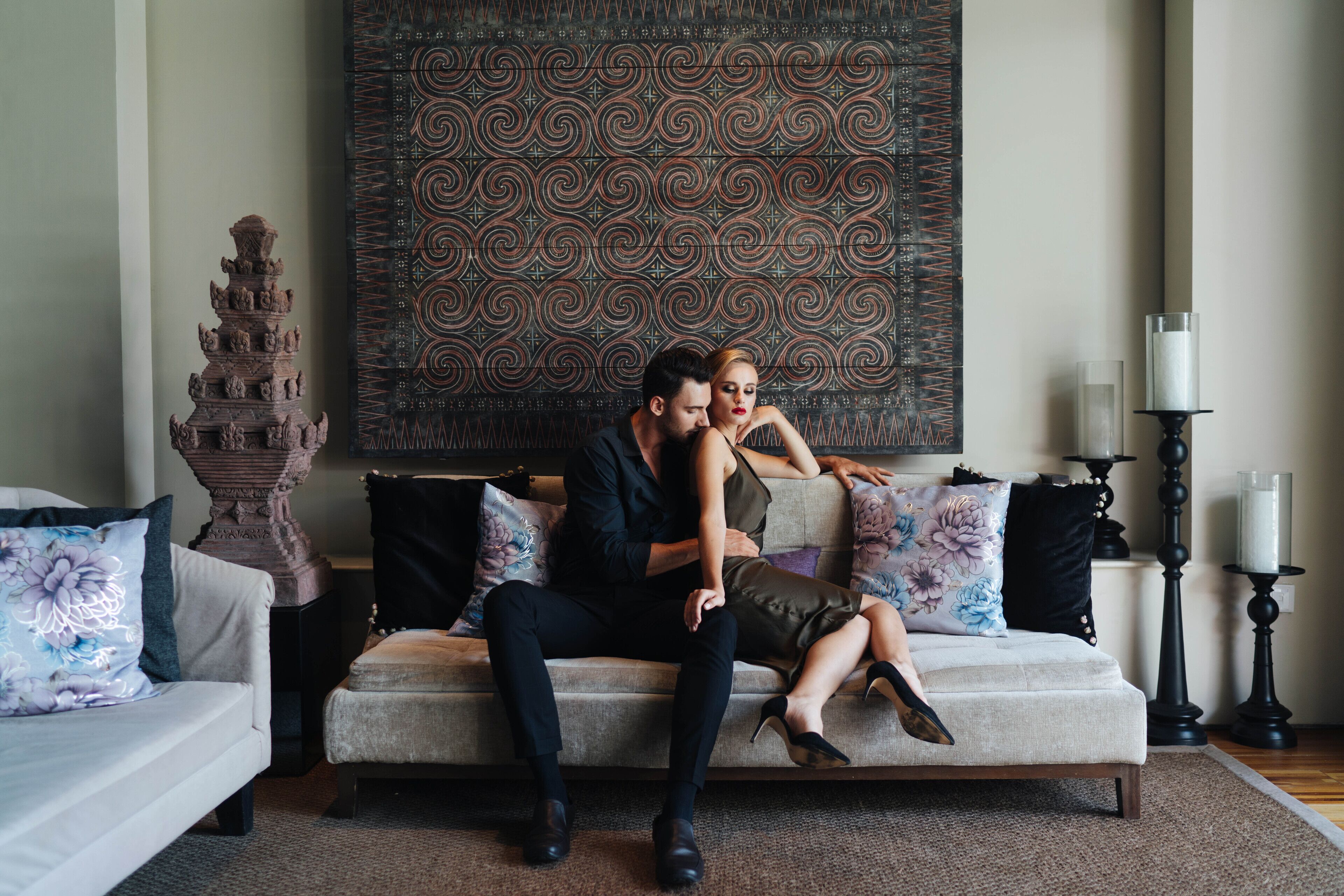 A couple sitting on a sofa with a patterned wall behind them, a tall vase, and a candle holder to the side.