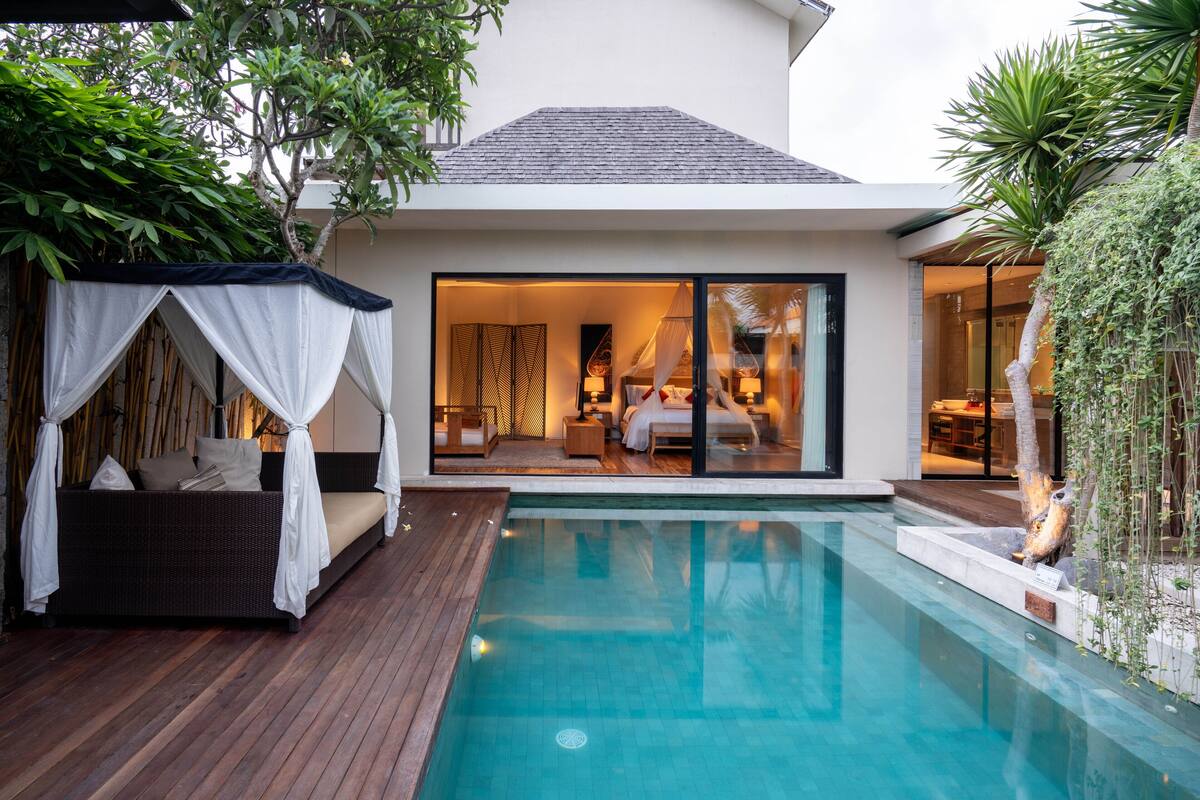 A modern outdoor pool area with a wooden deck, a wicker sofa with a canopy, and a glass door leading to a bedroom.