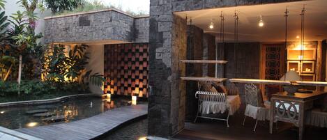 A modern outdoor dining area with a stone wall, wooden deck, and a small pond.