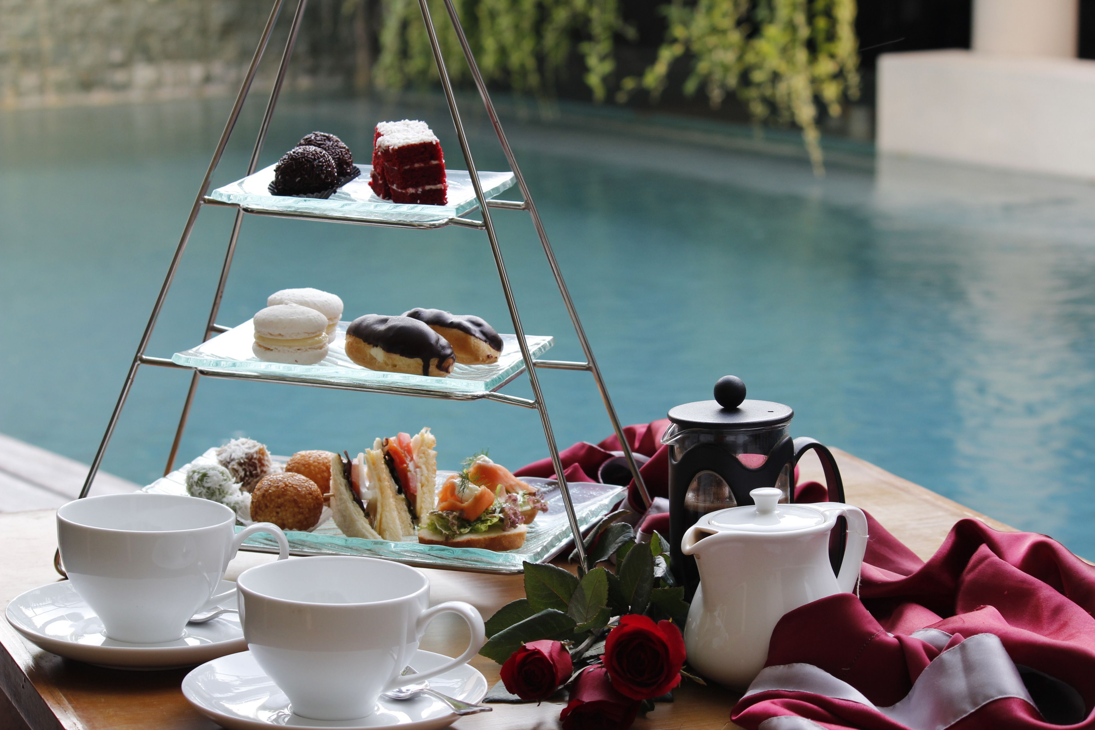 A tiered tray with pastries and sandwiches, a teapot, cups, and a red rose on a table by a pool.