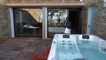 Uma banheira de hidromassagem numa área externa coberta, com um deck de madeira, paredes de pedra e uma porta de vidro que dá para um pátio.
