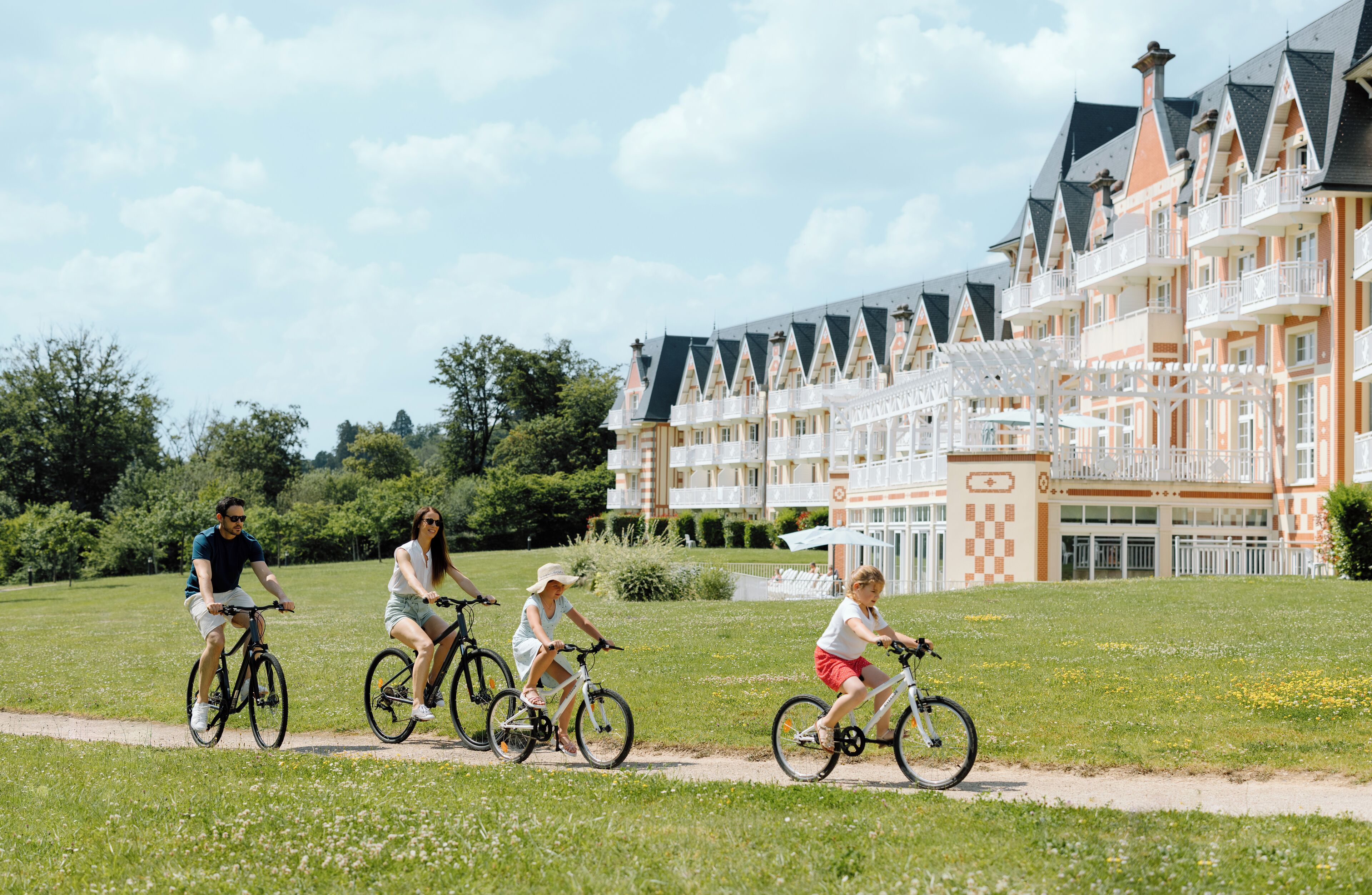 A group of people riding bicycles on a path in front of a large, ornate building with multiple balconies and a checkered pattern on the facade.