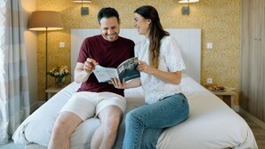 Una pareja sentada en una cama dentro de una habitación de hotel con papel tapiz estampado y dos lámparas de noche.