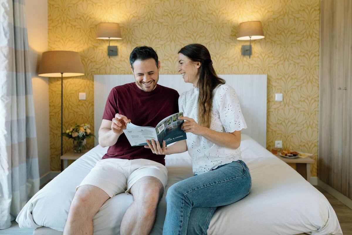 A couple sitting on a bed in a hotel room with patterned wallpaper and two bedside lamps.