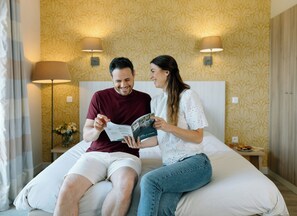 A couple sitting on a bed in a hotel room with patterned wallpaper and two bedside lamps.