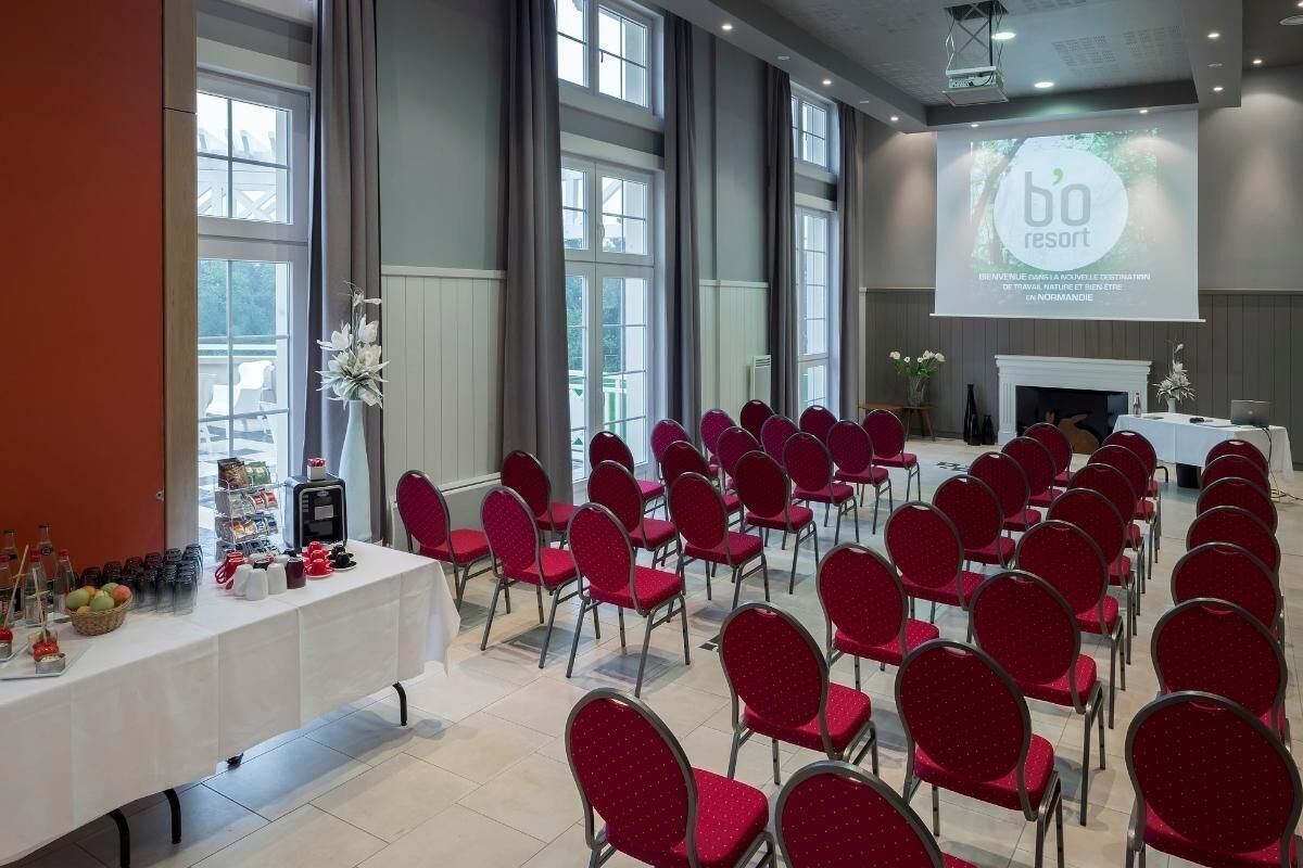 Een vergaderzaal met rijen rode stoelen, een witte tafel met diverse objecten, en een projectiescherm waarop 'bo resort' staat.