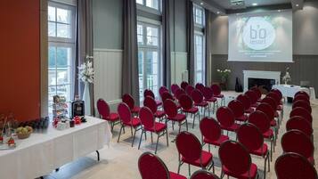 Een vergaderzaal met rijen rode stoelen, een witte tafel met diverse objecten, en een projectiescherm waarop 'bo resort' staat.