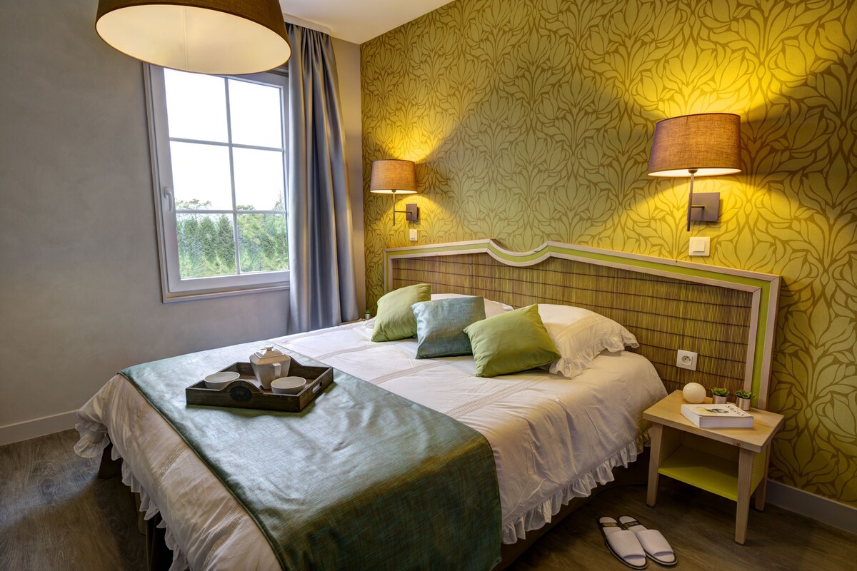 A hotel room with a bed, nightstand, lamp, window, and a tray with items on the bed.