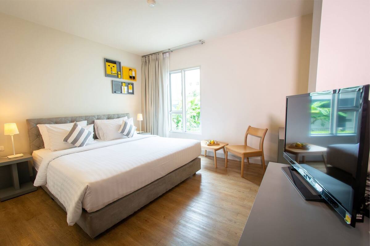 A hotel room with a bed, bedside tables, a TV, a chair, a small table, and a window with curtains.