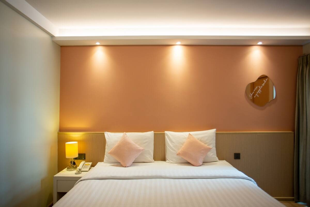A hotel room with a bed, a nightstand with a lamp and a phone, and a heart-shaped mirror on the wall.