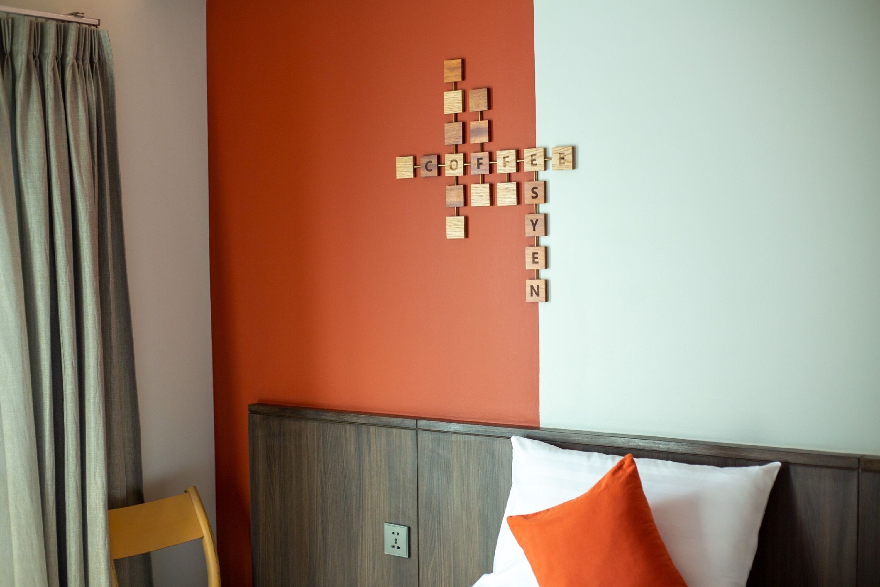 A room with a wooden headboard, a bed with white and orange pillows, and a wall with a crossword puzzle made of wooden blocks.
