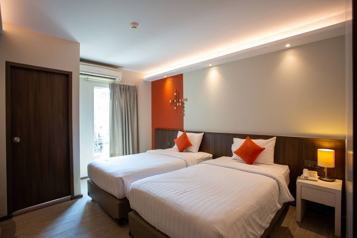 A hotel room with two beds, a nightstand with a lamp, a phone, and a view of the outside.
