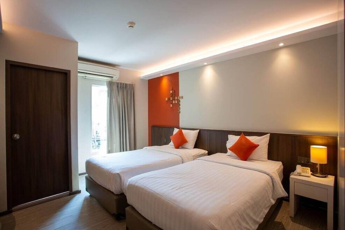 A hotel room with two beds, a wooden door, a nightstand with a lamp, and a window with curtains.