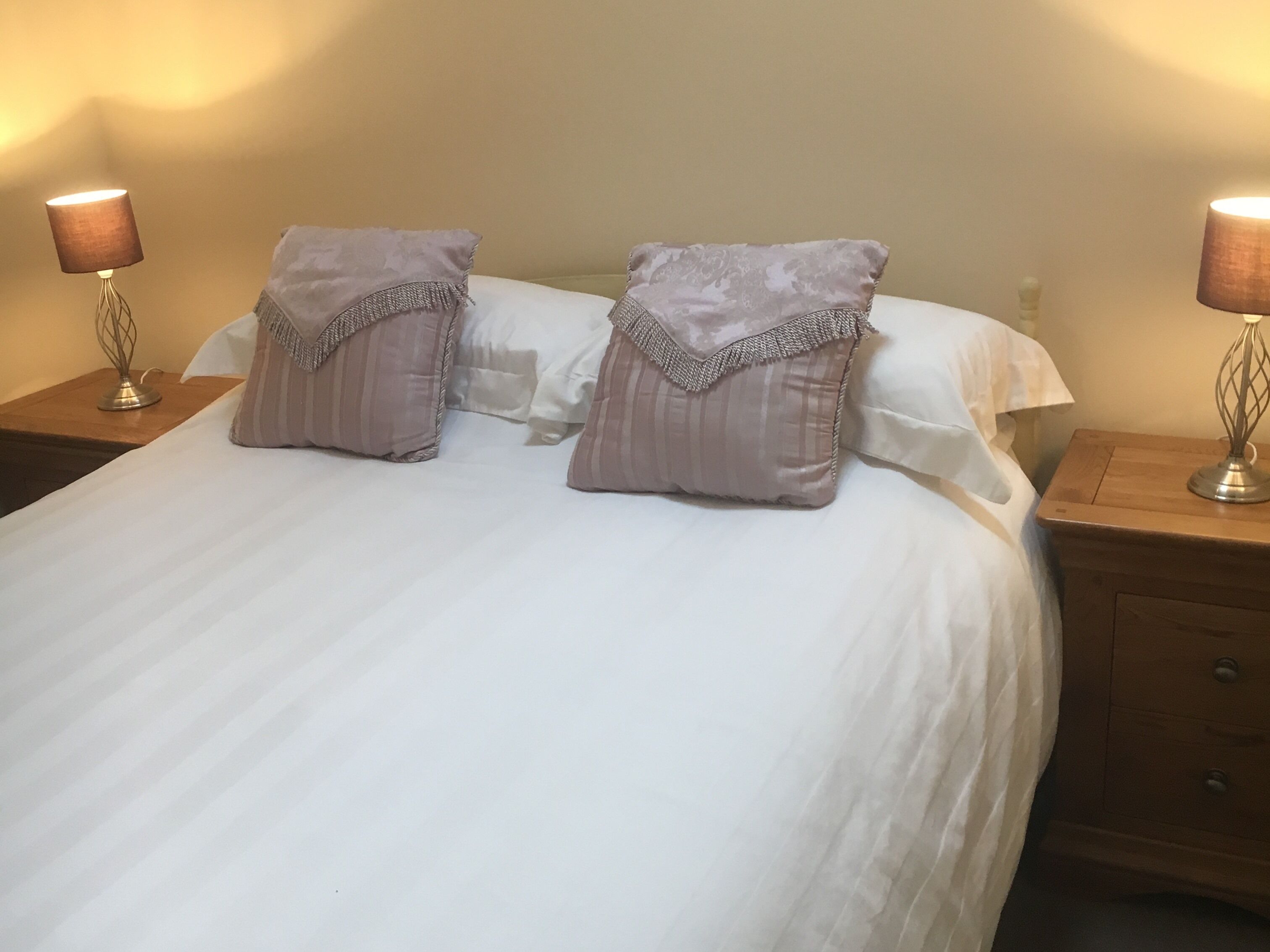 Una cama bien hecha con dos almohadas, una mesita de noche de madera con una lámpara y un cajón.