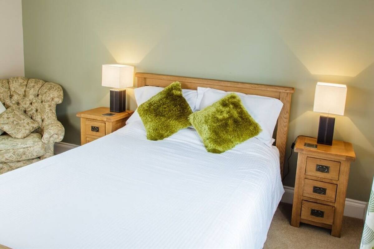 Una cama bien hecha con sábanas blancas y dos almohadas verdes, flanqueada por dos mesitas de noche con lámparas.