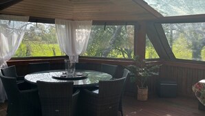 Un espacio al aire libre techado con una mesa redonda y sillas, una planta en maceta, y vistas a árboles y follaje.