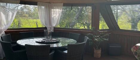 Un espace extérieur abrité, avec une table ronde et des chaises, un pot de fleurs et une vue sur les arbres et la verdure.