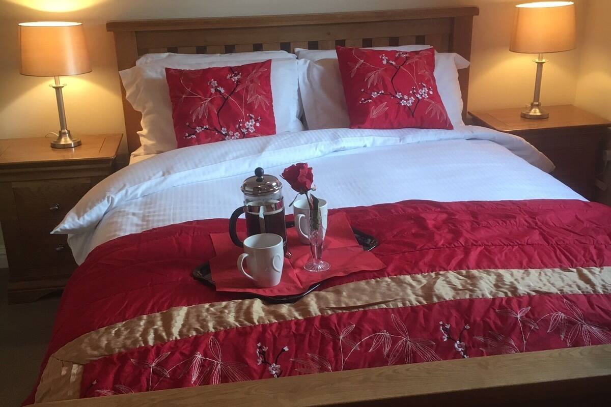 Una cama bien tendida con colcha roja y blanca, dos lámparas de noche y una mesita de noche con cafetera y taza.