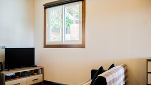 A room with a television on a wooden stand, a window with blinds, a bed with a striped blanket, and a small table with a lamp. - Gecko Lipe Resort (Koh Lipe)