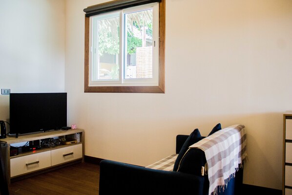 Una habitación con televisión sobre un soporte de madera, ventana con persianas, cama con manta a rayas y una pequeña mesa con lámpara.