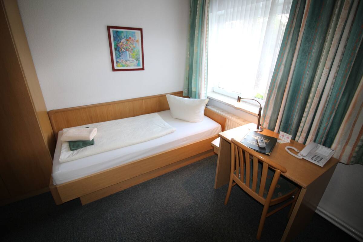 Ein Einzelbett mit Kissen und einem gefalteten Handtuch, ein Holztisch mit Telefon und ein Stuhl.