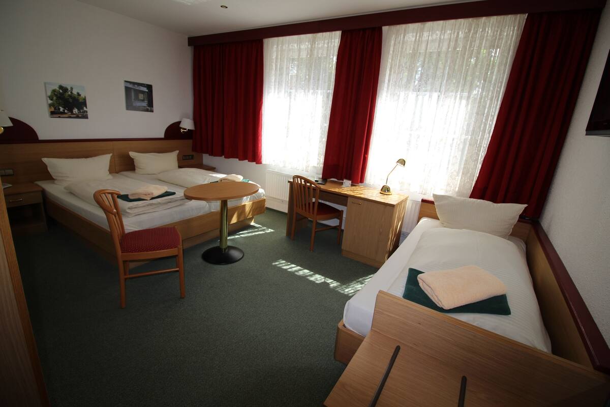 Ein Hotelzimmer mit zwei Betten, einem Schreibtisch und einem Stuhl. Zwei Fenster mit roten Vorhängen.