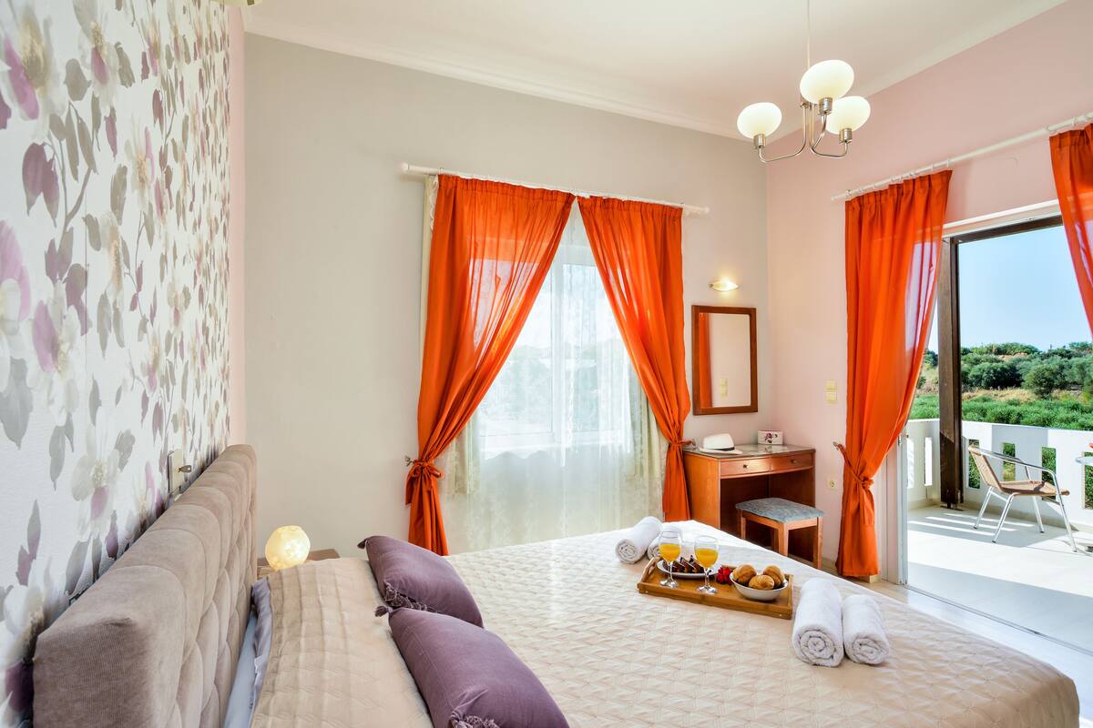 Habitación de hotel con cama, mesita de noche de madera, escritorio, silla y balcón con vistas.
