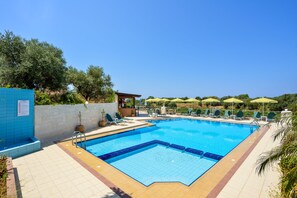 Piscina con sillas de descanso y sombrillas, rodeada por un suelo de baldosas y follaje.