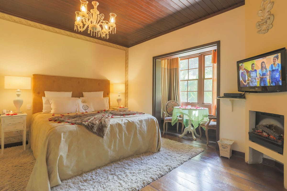 Un dormitorio con una cama grande, un candelabro, una chimenea y un televisor mostrando a un grupo de personas.