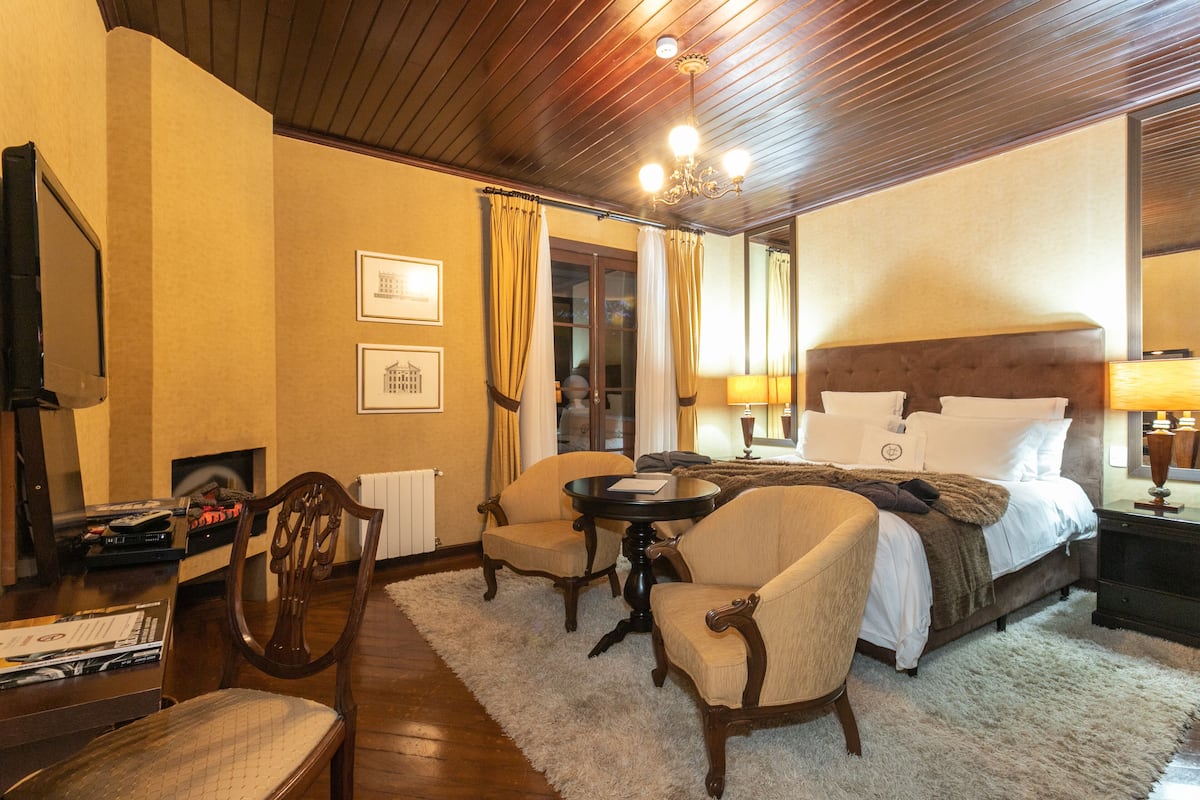 Una habitación de hotel con una cama, dos sillones, una mesita, una chimenea y cuadros enmarcados en la pared.