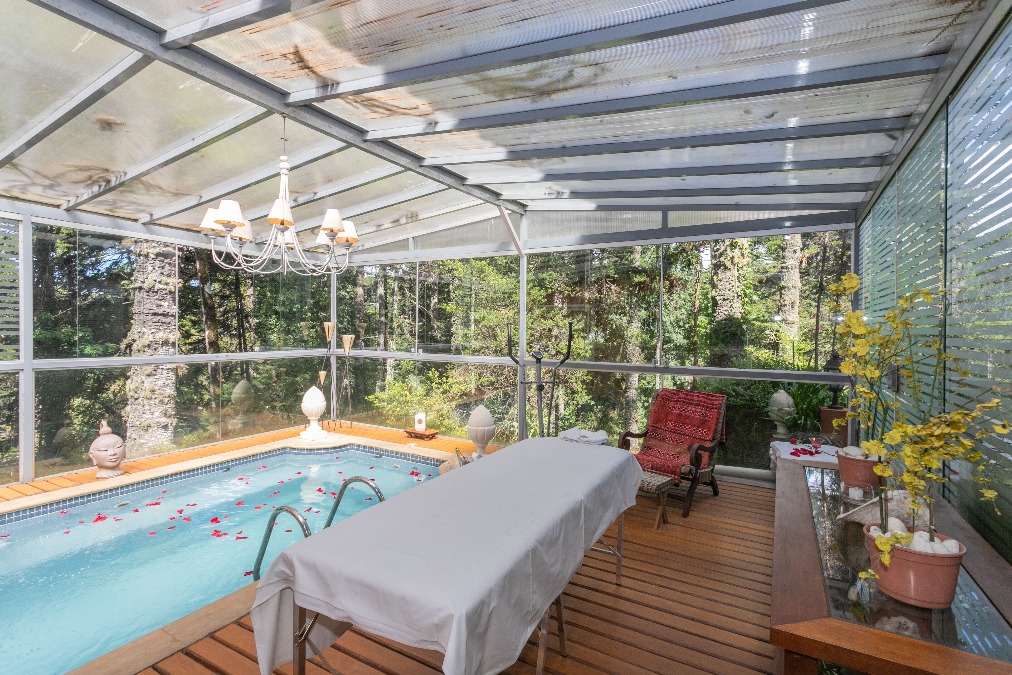 A glass-enclosed pool area with a wooden deck, a massage table, and a red chair.