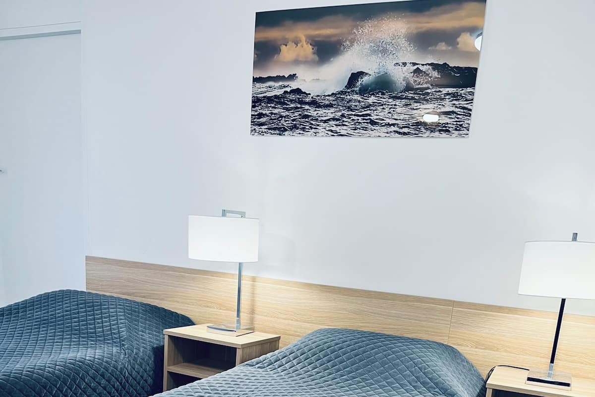 Une chambre d’hôtel avec deux lits, une tête de lit en bois, des tables de chevet avec des lampes et un tableau représentant un paysage côtier accroché au mur.
